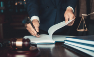 A lawyer writes in a notebook on a desk. A gavel and scales also sit on the desk.