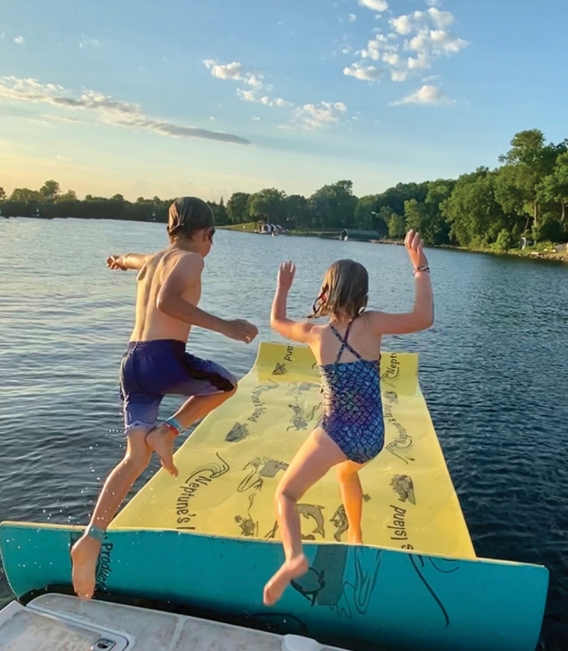 Two kids jumping onto a lily pad float in the lake.