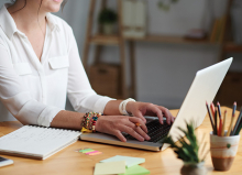 A woman sits working at a laptop with a pencil in hand and a notebook nearby.