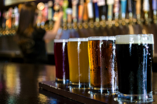 A flight of craft beers sits on a bar. Behind, a row of craft beers on tap.